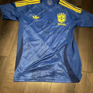 Blå Brasilien fotbollströja från Adidas - Säljer en blå Brasilien fotbollströja från Adidas med gula detaljer och tryck av Kristusstatyn på framsidan. Tröjan har korta ärmar, gula ränder på axlarna och Brasil-logga på bröstet. Slim fit-modell, tillverkad i Brasilien.