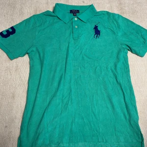 Turkos pikétröja från Polo Ralph Lauren - Säljer en turkos pikétröja från Polo Ralph Lauren med klassisk krage, knappar och stor marinblå logga broderad på bröstet. Siffra 3 i marinblått på ärmen. Perfekt för en sportig och stilren look. Denna är liten i storleken men hör av er om ni undrar något!