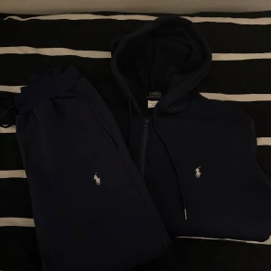 Mörkblå byxor och hoodie från Polo Ralph Lauren - Mörkblå byxor och hoodie från Polo Ralph Lauren med dragkedja och vit broderad logga på bröstet. Klassisk huva med dragsko och mjukt material. Perfekt för en avslappnad stil.