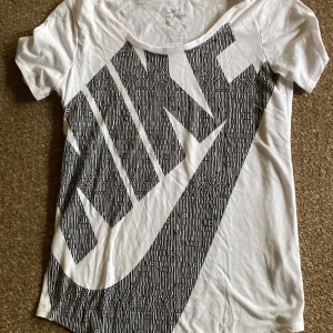 Vit t-shirt från Nike med stor logga - Vit t-shirt från Nike med stor svart logga och grafiskt mönster framtill. Klassisk rund halsringning och korta ärmar. Tillverkad i mjuk bomull för en bekväm känsla.