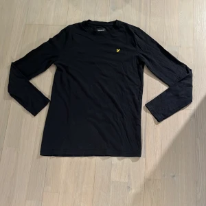 Svart långärmad tröja från Lyle & Scott - Svart långärmad tröja från Lyle & Scott med rund halsringning och den klassiska gula loggan broderad på bröstet. Tillverkad i mjuk bomull, perfekt för en stilren och enkel look. Den har tyvärr ett litet hål på högerbaksida. 