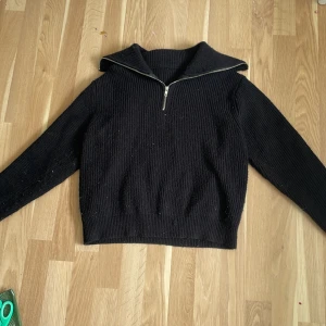 Svart stickad tröja med halv dragkedja - Svart stickad tröja med hög krage och halv dragkedja framtill. Tröjan har lång ärm och ribbad struktur, perfekt för kyligare dagar. Enkel och stilren design som passar till mycket.