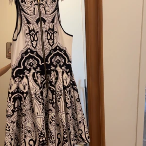 Vit och svart mönstrad klänning med blommor från Juicy Couture - Supersnygg ärmlös klänning från Juicy Couture med svart och vit grafisk design och färgglada blommor framtill. Klänningen har rund hals, dragkedja i ryggen och en utställd kjol som ger en feminin siluett.