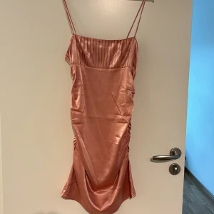 Rosa satinklänning med tunna axelband från NA-KD - Säljer en snygg rosa satinklänning från NA-KD med tunna axelband och veckad detalj upptill. Klänningen har en figurnära passform och är perfekt för sommarens tillfällen. Materialet är glansigt och ger en lyxig känsla.