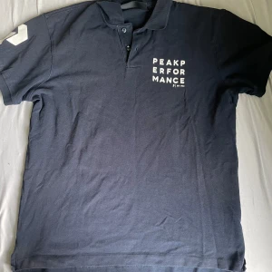 Mörkblå piké från Peak Performance - Säljer en mörkblå pikétröja från Peak Performance med vit logotyp på bröstet. Tröjan har korta ärmar, krage och knappar framtill. Tillverkad i 100% bomull för en skön känsla. Perfekt för en sportig och stilren look.