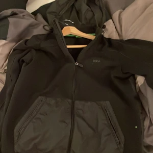 Svart hoodiejacka från BOSS - Svart hoodiejacka från BOSS med dragkedja. Jackan har en modern passform och diskret BOSS-logga på bröstet. Perfekt för en stilren och avslappnad look. Storlek L passar M