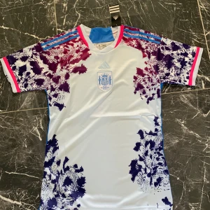 Vit och blå Spanien fotbollströja Adidas - Säljer en officiell Spanien fotbollströja från Adidas. Tröjan är vit med blå och lila mönster på axlar och sidor, samt röda detaljer. Har korta ärmar, blå Adidas-ränder på axlarna och Spaniens landslagslogga på bröstet.