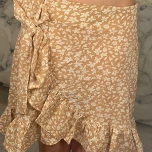 Rosa blommig kjol med volang från SHEIN - Supersöt rosa/beige kjol från SHEIN med vitt blommönster och volangkant. Kjolen har knyt i sidan och är kort, perfekt för varma dagar. Lätt och luftig känsla som ger en romantisk vibe.