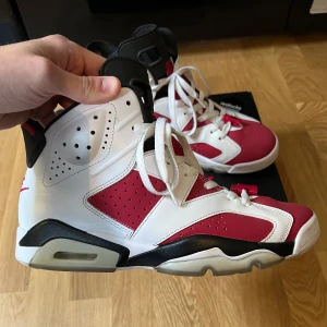 Nike Air Jordan 6 Retro 'Carmine' 2021 - Säljer ett par Nike Air Jordan 6 Retro 'Carmine' 2021 med vit och röd ovandel, svarta detaljer och synlig Air-sula. Klassisk siluett med snörning och rund tå. Perfekta för dig som gillar sneakers med ikonisk design. Box och tag från restocks medföljer. Använda 3 gånger 8/10 i skicket!