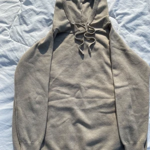 Beige stickad hoodie med snörning - Säljer en beige stickad hoodie med huva och snörning framtill. Tröjan har långa ärmar och ribbade muddar vid ärmslut och nederkant. Perfekt för en avslappnad och stilren look.