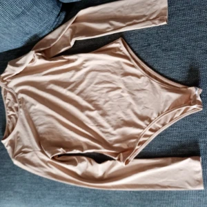 Beige långärmad bodysuit från SHEIN stl M - Säljer en beige långärmad bodysuit från SHEIN. Den har rund halsringning och är tillverkad i ett mjukt, stretchigt material som sitter tight mot kroppen. Perfekt att styla med jeans eller kjol.