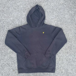 Svart hoodie från Lyle & Scott Junior - Säljer en svart hoodie från Lyle & Scott Junior med klassisk gul logga på bröstet. Tröjan har huva, magficka och långa ärmar. Perfekt för en avslappnad stil och enkel att matcha med det mesta.