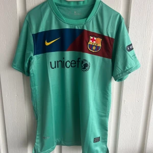 FC Barcelona fotbollströja Messi 10 - Snygg FC Barcelona fotbollströja i mintgrönt med klubbmärke, Nike-logga och UNICEF-tryck på framsidan. Messi 10 och Champions League-märke på ärmen. Kortärmad modell i lätt och ventilerande material.