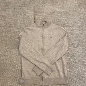 Beige kofta från Morris - Säljer en stilren ljusgrå kofta från Morris i 100% merinoull. Tröjan har dragkedja framtill, hög krage och diskret logga på bröstet. Perfekt för lager-på-lager och passar till många olika stilar.