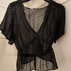 Zara mesh blus - Säljer denna fina svarta transparent omlottblus med korta ärmar och små prickar i tyget. Blusen har spetsdetaljer vid halsringningen och resor i midjan. Perfekt för sommaren och knappt använd! 💝