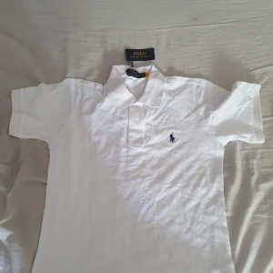 Vit piké t-shirt från Polo Ralph Lauren - Vit piké t-shirt från Polo Ralph Lauren med klassisk krage och två knappar framtill. Broderad marinblå logga på bröstet. Tillverkad i bomull och har en slim fit passform.