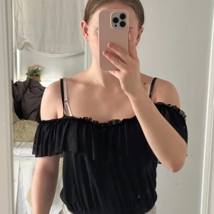 Svart offshoulder body med volang - Svart body med offshoulder och volangdetalj runt bysten. Använd enstaka gånger! Står ingen storlek men skulle gissa på S.