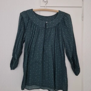 Mörkgrön blus med blommigt mönster från Nygårdsanna - Säljer en mörkgrön blus från Nygårdsanna med diskret blommigt mönster. Blusen har trekvartsärm, rund halsringning och en knapp framtill. Tillverkad i 100% bomull vilket gör den mjuk och skön att bära.
