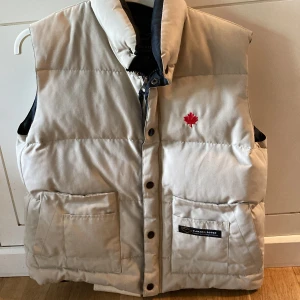 Beige dunväst från Canada Goose - Säljer en beige dunväst från Canada Goose med röd lönnlöv-detalj på bröstet och två stora fickor framtill. Västen har hög krage, knappar och dragkedja samt svart innerfoder. Perfekt för lager-på-lager under kyliga dagar.