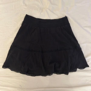 Svart kjol från Gina Tricot - Svart kjol från Gina Tricot med bred resår i midjan och dekorativa spetsdetaljer. Kjolen har en lätt utställd form och är tillverkad i ett tunt, mjukt tyg som ger ett flowy intryck. Perfekt att styla med en enkel topp eller stickad tröja.