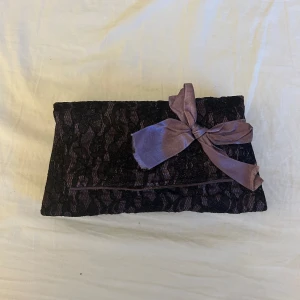 Svart spets kuvertväska med lila rosett - Säljer en svart kuvertväska med spetsdetaljer och en stor lila rosett framtill från Graceland. Väskan har avtagbar kedja så den kan bäras både i handen och över axeln. 