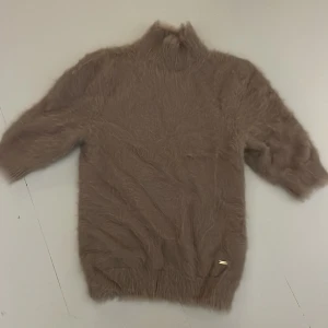 Beige stickad T-shirt från Lindex - Mysig beige stickad tshirt från Holly & White by Lindex. Tröjan är fluffig och har kort ärm samt hög krage. Perfekt för kyliga dagar när du vill vara både varm och snygg.