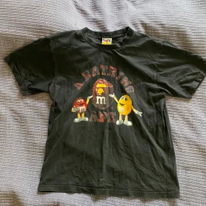 Svart M&M's x BAPE t-shirt - Svart t-shirt från A Bathing Ape med färgglatt M&M's-tryck framtill. Klassisk passform med rund hals och korta ärmar.så klart så rollar jag av håret tvättar och stryker plagget innan jag skickar, ifall någon är katt allergisk.  Står att den är Large men passar som en M, fråga gärna efter mått.