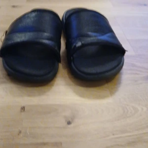 Svarta sandaler med bred rem - Säljer ett par svarta sandaler med bred rem över foten. De har en enkel och stilren design med platt sula och öppen tå, perfekta för varma dagar. Materialet känns syntetiskt och de är lätta att ta på och av.