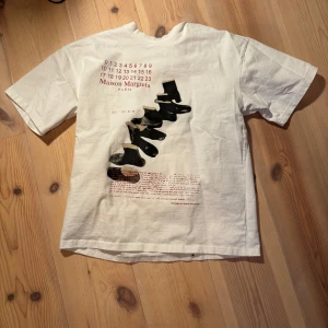 Vit t-shirt från Maison Margiela passar L och XL - Vit t-shirt från Maison Margiela med unikt svart och rött tryck på framsidan. Klassisk passform och rund halsringning. Perfekt för dig som gillar statement-plagg och exklusiva märken.