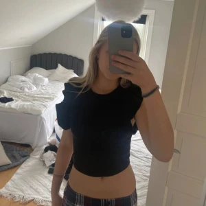 Svart croppad topp med volangärmar - Säljer en svart croppad topp med volangdetaljer på ärmarna. Toppen har en tight passform och är perfekt för dig som vill ha en trendig och stilren look. Passar till både jeans och kjol.