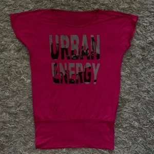 Mörkrosa t-shirt med tryck Urban Energy - Cool mörkrosa t-shirt med svart och grått tryck där det står 'URBAN ENERGY' och detaljer av små glittriga stenar på bokstäverna. T-shirten har korta ärmar och en lös passform, perfekt för en avslappnad streetstil.