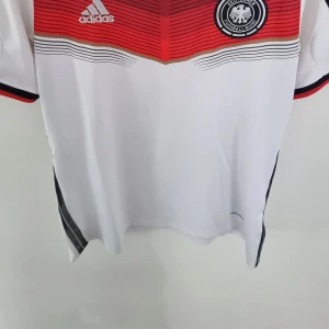 Tysklands fotbollströja Adidas 2014 VM - Säljer en officiell Tyskland fotbollströja från Adidas. Tröjan är vit med svarta, röda och guld detaljer samt tunna svarta ränder upptill. Kortärmad modell med broderad landslagsemblem och Adidas-logga på bröstet. Perfekt för fotbollsfans! Från 2014 VM Storlek S (herr)