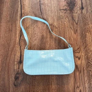Ljusblå axelväska i syntet - Säljer en stilren ljusblå axelväska med rutmönstrad yta. Väskan har en justerbar axelrem och en enkel, modern design som passar perfekt till olika outfits. 