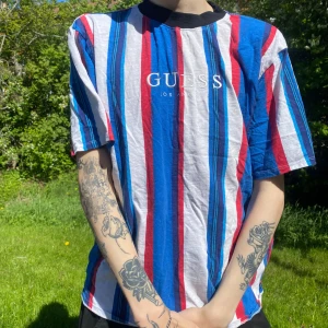 Randig t-shirt från Guess Originals - Säljer en färgglad randig t-shirt från Guess Originals i storlek L. T-shirten har breda vertikala ränder i blått, rött, vitt och svart samt logga på bröstet. Påminner om klubbor och tivolin.