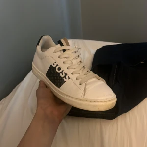 Vita sneakers från BOSS med svart detalj - Snygga vita sneakers från BOSS med svart logga på sidan och svarta detaljer vid hälen. Skorna har snörning och en platt sula, perfekt för en clean och stilren look. Klassisk rund tå och diskret design.