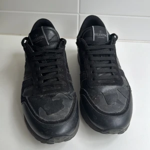 Valentino Garavani Rockrunner sneakers i svart - Snygga svarta Valentino Garavani Rockrunner sneakers med diskret camouflage-mönster. Skorna har snörning, rund tå och platt sula. Materialmix av mocka och skinn ger en lyxig känsla. Perfekta för dig som vill ha en stilren och exklusiv look.