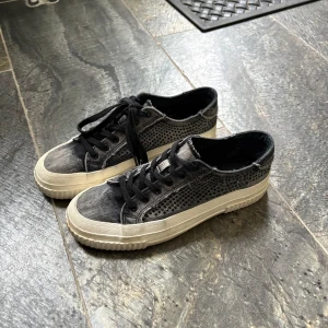 Svarta sneakers med vit sula - Säljer ett par svarta sneakers med vit, räfflad sula och svarta skosnören. Skorna har ett coolt perforerat mönster på sidorna och en något sliten look vid kanterna. Perfekta för dig som gillar en avslappnad och trendig stil.