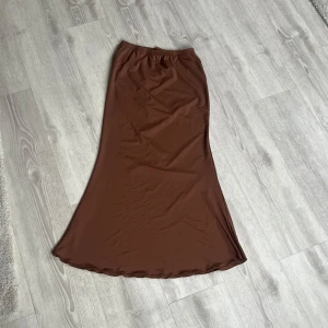 Brun långkjol i satin - Säljer en brun långkjol i satin med enkel och stilren design. Kjolen har resår i midjan och ett lätt utsvängd passform som ger en snygg siluett. Perfekt att styla med en enkel topp eller stickad tröja. 