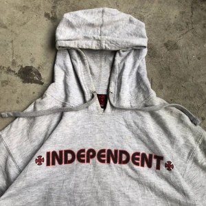 Grå hoodie från Independent Truck Co - Säljer en grå hoodie från Independent Truck Company med stor röd och svart logga på ryggen och texten 'INDEPENDENT' framtill. Tröjan har huva med snörning och är tillverkad i en mjuk bomulls- och polyestermix. Perfekt för dig som gillar streetwear och skate.