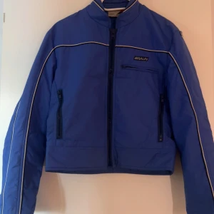 Blå vindjacka från Braun med vita detaljer - Snygg blå vindjacka från Braun med vita piping-detaljer längs ärmar och bröst. Jackan har hög krage, dragkedja framtill och flera praktiska fickor med dragkedja. Perfekt för dig som gillar sportig och clean stil.