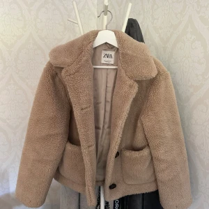 Beige teddyjacka från Zara XS - Supermjuk och fluffig teddyjacka från Zara i beige färg. Jackan har stora fickor framtill, bred krage och stängs med knappar. Perfekt för dig som vill ha en mysig och trendig look under kyliga dagar.