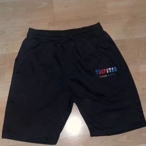  Trapstar shorts - Svarta shorts från Trapstar med broderad logga i blått, rött och vitt på vänster ben. Mjuk bomullskänsla och elastisk midja med snörning för skön passform. Perfekta för chill dagar eller när du vill ha en avslappnad streetstyle.
