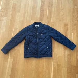 Mörkblå quiltad jacka från Holly & Whyte - Quiltad mörkblå jacka från Holly & Whyte med snygga detaljer som dragkedja framtill, fickor med tryckknappar och en extra bröstficka med zip. Jackan har långärmad passform och står ut med sitt stilrena rutmönster. Tveka inte att kontakta vid frågor.