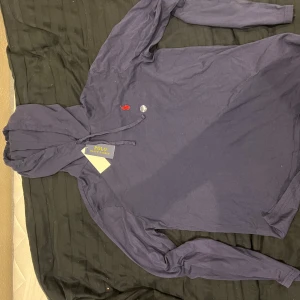 Marinblå hoodie från Polo Ralph Lauren - Snygg och helt ny tunn marinblå hoodie från Polo Ralph Lauren med klassisk röd logobrodyr på bröstet. Tröjan har huva med snörning och långa ärmar. Tillverkad i mjuk bomull som känns skön mot huden. Lappen kvar