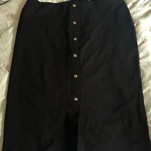 Svart långkjol med knappar från H&M - Stilren svart långkjol från H&M i storlek XXL. Kjolen har raka linjer och snygga guldfärgade knappar framtill som ger en clean look. Perfekt att matcha med en crop top eller stickad tröja. Materialet känns mjukt och följsamt.