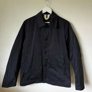 Mörkblå jacka från Arket, stl 46 - Klassisk mörkblå jacka från Arket i stilren design. Jackan har rak passform, krage, knäppning med svarta knappar och två sidofickor. Tillverkad i kompakt polyesterblandning, perfekt för dig som gillar minimalistisk och clean look. Px0e