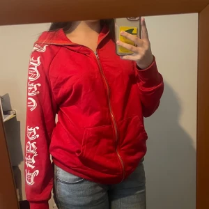 Röd hoodie med tryck på ärmen - Säljer en röd hoodie med dragkedja och huva. Den har coolt vitt tryck längs ena ärmen och två fickor framtill. Hoodien kan stängas hela vägen (luvan också).
