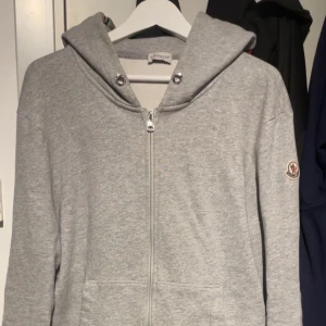Grå Moncler hoodie med dragkedja - Snygg grå hoodie från Moncler med huva och silverfärgad dragkedja framtill. Tröjan har Moncler-logga på vänster ärm och två fickor fram. Tillverkad i mjuk bomull, perfekt för chill dagar. Stilsäker och enkel att matcha med jeans eller joggers. Snören kan du få med och är jätte enkelt att sätta tillbaka! Pris kan diskuteras. 
