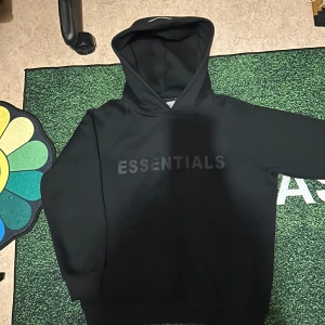 Svart Essentials Fear of God hoodie - Svart hoodie från Essentials Fear of God med tryckt logga på bröstet och ärmen. Klassisk oversized passform, stor huva och ribbade muddar. Perfekt för en clean och stilren streetwear-look.