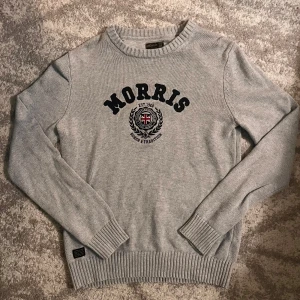 Grå stickad tröja från Morris - Snygg grå stickad tröja från Morris med svart broderad logga och emblem på bröstet. Tröjan har rund hals, ribbade muddar och nederkant samt lång ärm. Perfekt för dig som gillar klassisk och stilren look.